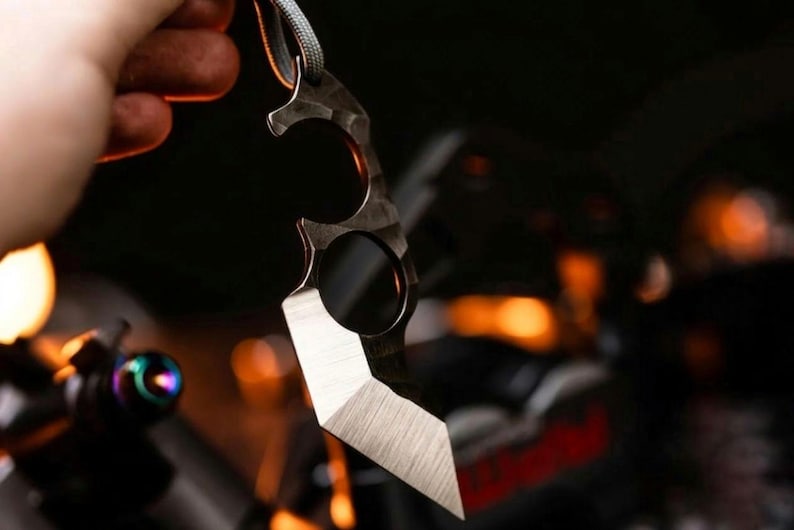 May include: A dark metal karambit knife with a textured finish and a lanyard. The blade has a sharp, angled edge. The knife is held against a blurred background with hints of orange and black.
