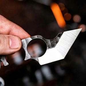 May include: A small, silver utility knife with a finger hole and a sharp, angled blade. The knife has a textured surface and is held by a hand. A grey lanyard is attached to the knife.