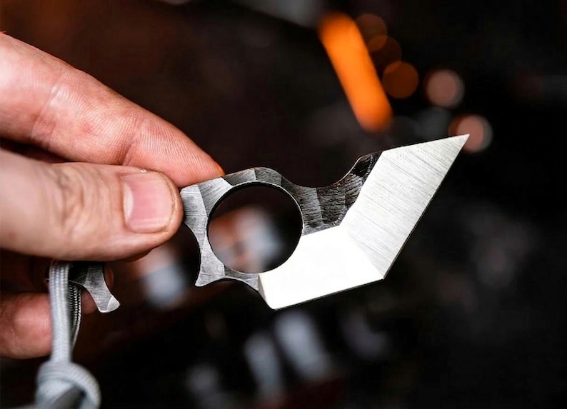 May include: A small, silver utility knife with a finger hole and a sharp, angled blade. The knife has a textured surface and is held by a hand. A grey lanyard is attached to the knife.