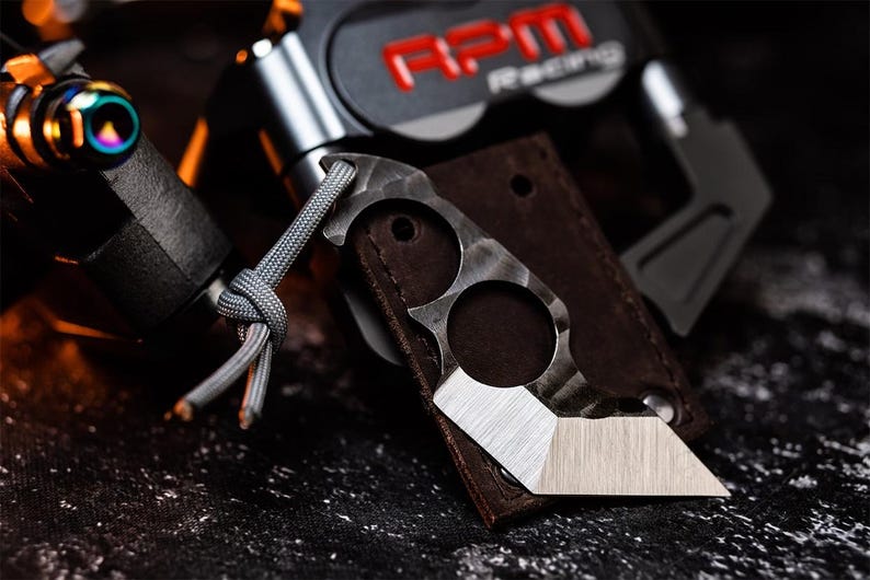 May include: A small, silver and black pocket knife with a textured blade and two finger holes. The knife is attached to a brown leather sheath with a grey cord. The background includes a black object and the text "RPM Racing".
