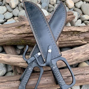 Puede incluir: Dos cuchillos sai negros con fundas de cuero. Los cuchillos tienen mangos texturizados y una hoja curva. Las fundas son de cuero negro con costuras decorativas. Los cuchillos descansan sobre un lecho de rocas y madera flotante.