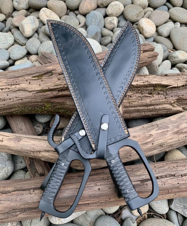 Puede incluir: Dos cuchillos sai negros con fundas de cuero. Los cuchillos tienen mangos texturizados y una hoja curva. Las fundas son de cuero negro con costuras decorativas. Los cuchillos descansan sobre un lecho de rocas y madera flotante.