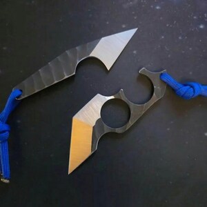 May include: Two small, handmade metal knives with sharp, angled blades. The blades have a silver finish, while the handles have a textured, dark grey appearance. Each knife is attached to a blue cord. The knives are displayed on a dark surface.