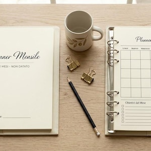 Può includere: Due agende ad anelli color crema con testo nero, incluse le parole Planner Mensile, 12 Mesi - Non Datato e Luglio. Una tazza in ceramica bianca con un disegno di foglie dorate, due clip metalliche dorate e una matita nera sono poste tra loro.