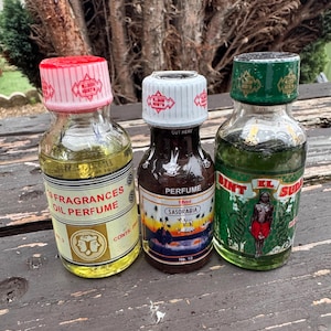 Puede incluir: Tres pequeños frascos de perfume de vidrio con etiquetas y tapones coloridos. Los frascos contienen líquidos amarillos, marrones y verdes. Las etiquetas presentan texto e ilustraciones, incluidas las palabras "FRAGRANCES OIL PERFUME" y "PERFUME SASORABIA".