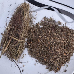 Può includere: Un fascio di erbe essiccate legato con spago accanto a un mucchio di foglie e steli secchi sfusi. Le erbe sono marroni e beige, e il piatto è bianco con accenti neri e grigi.