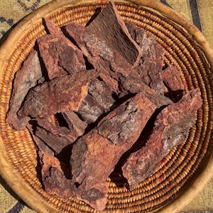 Puede incluir: Una cesta de mimbre llena de trozos de corteza de árbol de color marrón rojizo. La corteza tiene una superficie rugosa y texturizada con tonos de marrón y rojo. La cesta está hecha de un material tejido marrón claro, y el fondo es una tela a rayas amarillas y azules.