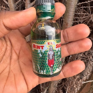Puede incluir: Pequeña botella de vidrio transparente con una etiqueta verde y dorada que presenta una figura con falda roja. La botella tiene una tapa verde y contiene un líquido oscuro. El texto "BINT EL SUD" es visible.
