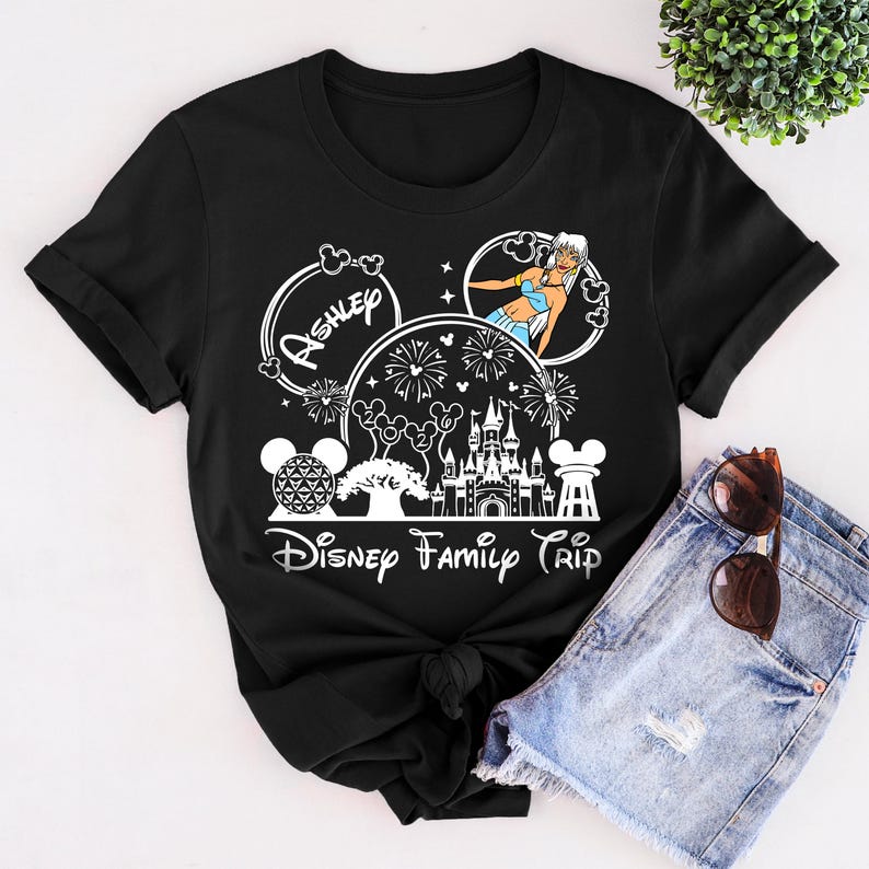 Puede incluir: Camiseta negra con un dise&ntilde;o gr&aacute;fico blanco con tem&aacute;tica de Disney. El dise&ntilde;o incluye el nombre "Ashley", un castillo, fuegos artificiales y el texto "Disney Family Trip".
