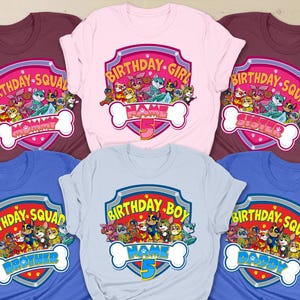 Può includere: Diverse magliette colorate con un design a cartoni animati e la scritta "BIRTHDAY SQUAD". Le magliette sono in vari colori, tra cui bordeaux, rosa e blu. Ogni maglietta ha un titolo diverso, come "Birthday Girl" o "Birthday Boy".