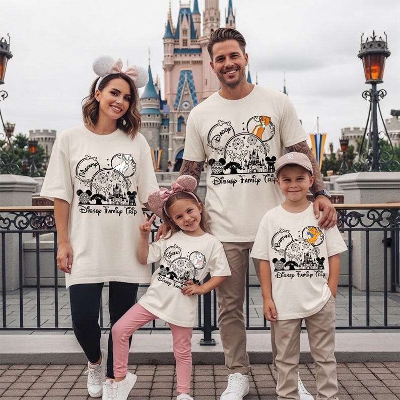Puede incluir: Cuatro personas vestidas con camisetas crema a juego con un dise&ntilde;o tem&aacute;tico de Disney. El dise&ntilde;o incluye orejas de Mickey Mouse, un castillo y el texto "Disney Family Trip". La familia posa frente a un castillo.