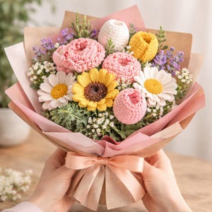 以下が含まれることがあります： ピンクのバラ、黄色のヒマワリ、白いデイジーなど、手作りのかぎ針編みの花束。茶色とピンクの紙で包まれています。 桃色のリボンで結ばれています。 花は緑に囲まれています。