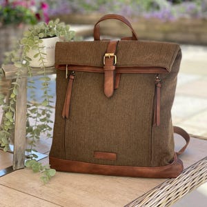 May include: A brown backpack with a flap closure and leather detailing. The bag has a top handle, adjustable straps, and a buckle fastening. The backpack is made of a textured fabric with leather trim and a small leather tag at the base.