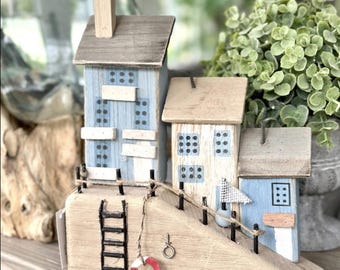 May include: A decorative wooden beach scene featuring miniature houses in shades of blue and white. The houses have small windows and are connected by a wooden ramp with a ladder, a life preserver, and a small flag. The scene is set on a wooden base.