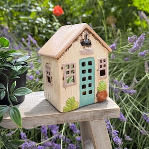 Puede incluir: Una casa de cerámica decorativa con exterior color crema, puerta de entrada color verde azulado y marcos de ventana rosados. La estructura presenta un techo texturizado, una pequeña lámpara negra sobre la puerta y follaje verde en la base.