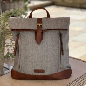 May include: A grey herringbone backpack with brown leather detailing on the base, straps, and buckle. It has a top handle and two side zip pockets. The bag's design is casual and stylish, suitable for everyday use.