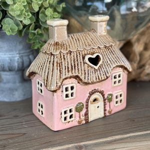 May include: A pink ceramic cottage with a thatched roof and heart-shaped window. The cottage has a white door, small windows, and two chimneys. Decorative potted trees flank the door. The cottage is a decorative item.