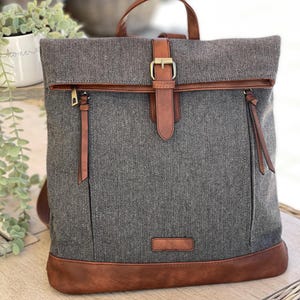 May include: Grey herringbone backpack with brown leather detailing. It has a top handle, a buckle closure, and two zipped pockets. The base and trim are brown leather. A fashionable and practical bag.