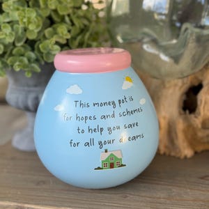 Peut inclure: Un pot à argent bleu clair avec un bord rose, avec une illustration de maison et le texte : « Ce pot à argent est pour les espoirs et les projets pour vous aider à économiser pour tous vos rêves. »