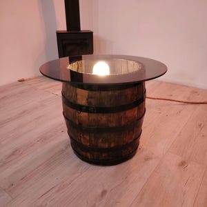 Puede incluir: Una mesa rústica de barril con una tapa redonda de cristal transparente. El barril de madera está sujeto con bandas metálicas oscuras. Una bombilla es visible dentro del barril, iluminando la tapa de cristal. La mesa mide aproximadamente 61 cm de altura.