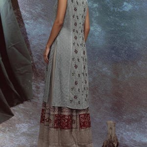 Puede incluir: Un vestido sin mangas de color gris claro con bordados florales y una abertura lateral. El vestido est&aacute; superpuesto a una falda beige con un dobladillo estampado en rojo y blanco. La modelo est&aacute; de pie frente a un fondo texturizado.