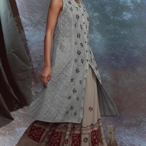 Puede incluir: Una t&uacute;nica sin mangas de color gris claro con bordados florales y una abertura alta, combinada con una falda beige con paneles estampados en rojo y blanco. El conjunto forma parte de una colecci&oacute;n de inspiraci&oacute;n bohemia.