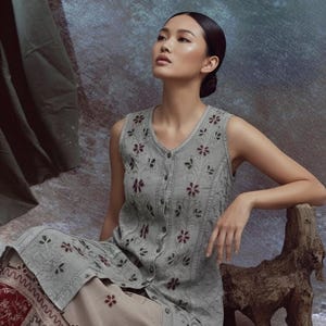 Puede incluir: Un vestido sin mangas, abotonado, de color gris claro con bordados florales y una falda a juego. La falda tiene un borde decorativo con estampado rojo y blanco. La modelo est&aacute; sentada junto a una escultura de madera.