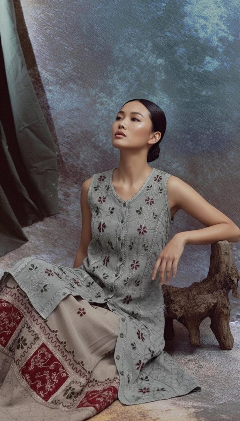 Puede incluir: Un vestido sin mangas, abotonado, de color gris claro con bordados florales y una falda a juego. La falda tiene un borde decorativo con estampado rojo y blanco. La modelo est&aacute; sentada junto a una escultura de madera.