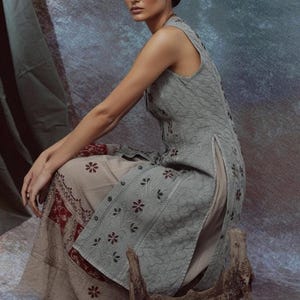 Puede incluir: Una mujer con una t&uacute;nica sin mangas verde claro y pantalones a juego con bordados florales. El conjunto tiene una abertura lateral y se combina con una falda beige con un borde estampado en rojo y blanco. Una escultura de madera est&aacute; en primer plano.