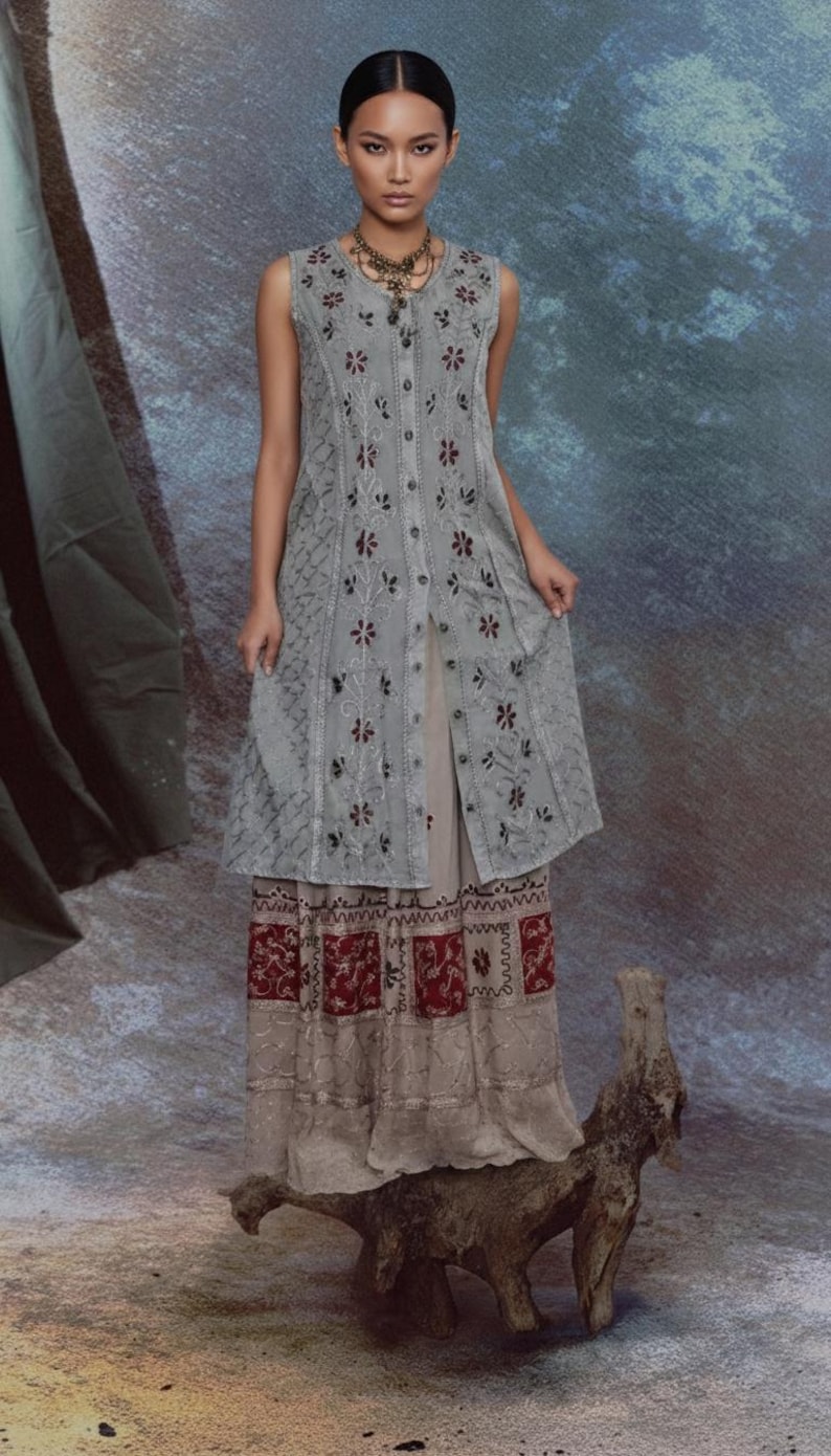 Puede incluir: Un vestido gris sin mangas con bordado floral y botones en la parte delantera. El vestido est&aacute; superpuesto a una falda beige con un dobladillo estampado en rojo y blanco. Se lleva un collar alrededor del cuello.