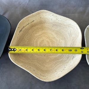 May include: A beige, irregularly shaped bowl with a light brown, swirled pattern. The bowl is approximately 12 inches long. A yellow measuring tape is placed across the bowl.