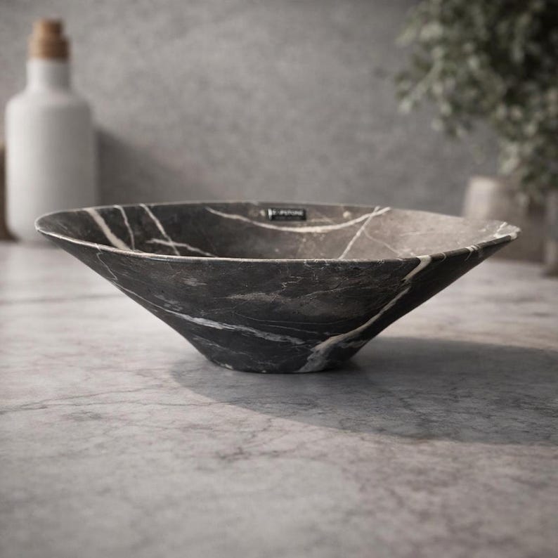 May include: A dark gray, marble-patterned bowl with white veining. The bowl has a flared, angular shape and sits on a gray surface. The bowl is likely for decorative or functional use in a bathroom or kitchen.