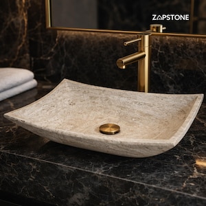 May include: A rectangular beige stone vessel sink with a textured surface and a centered gold drain plug. A tall gold faucet is positioned behind the sink. The text ZAPSTONE is visible in the upper right corner of the image.