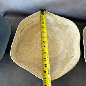 May include: A beige, irregularly shaped bowl with a light brown, marbled pattern. The bowl is approximately 12 inches in length, as measured by a yellow tape measure. The bowl is next to a dark blue bowl.