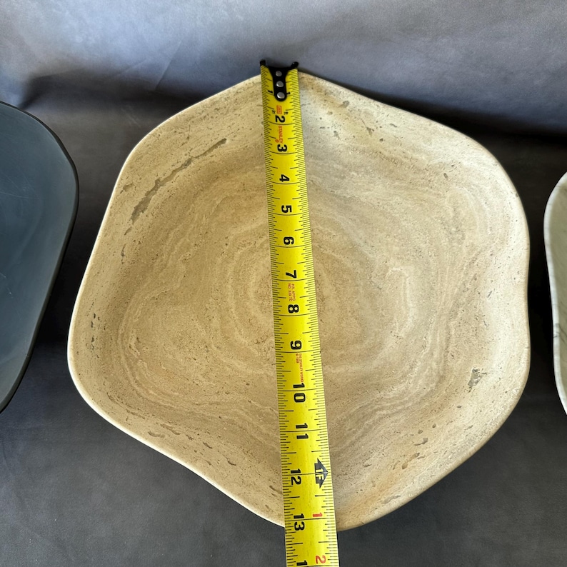 May include: A beige, irregularly shaped bowl with a light brown, marbled pattern. The bowl is approximately 12 inches in length, as measured by a yellow tape measure. The bowl is next to a dark blue bowl.