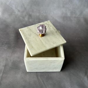 May include: A square, off-white marble box with a lid. The lid is slightly ajar, revealing the interior. A light purple crystal knob sits atop the lid. The box is a decorative item, suitable for storing small items.