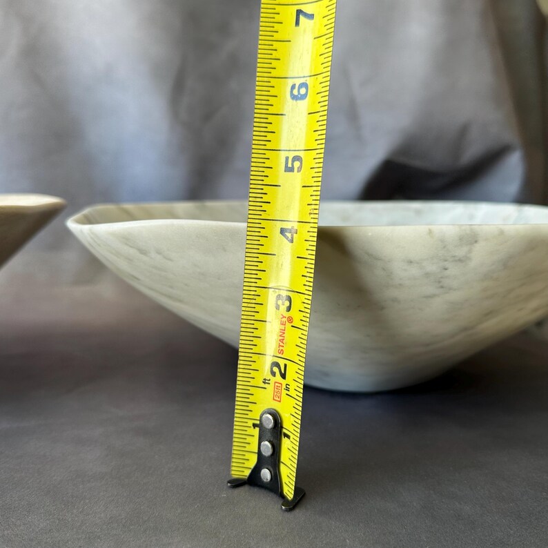 May include: A light-colored, marbled bowl with a geometric shape. The bowl is approximately 3 inches (7.6 cm) deep. A yellow tape measure is positioned next to the bowl, showing the depth.