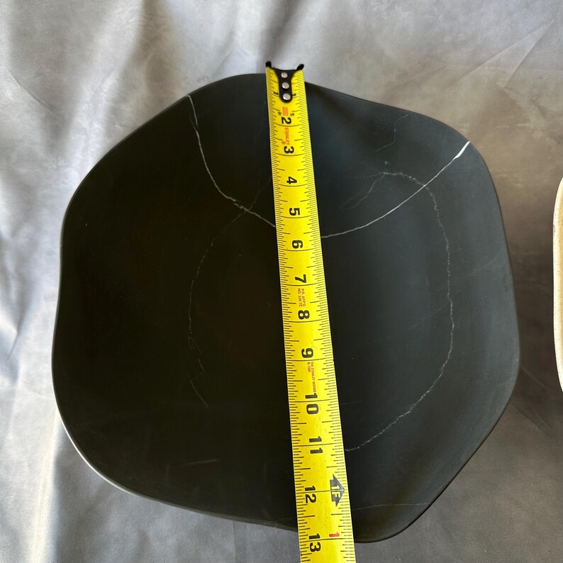 May include: A black, irregularly shaped serving dish with white veining. The dish is approximately 12 inches long, as measured by a yellow tape measure. The dish has a matte finish and a unique, organic form.