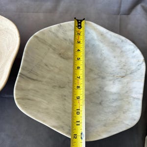 May include: A white marble serving dish with gray veining. The dish is irregularly shaped and measures approximately 11 inches in length. A yellow measuring tape is placed next to the dish to show its size.