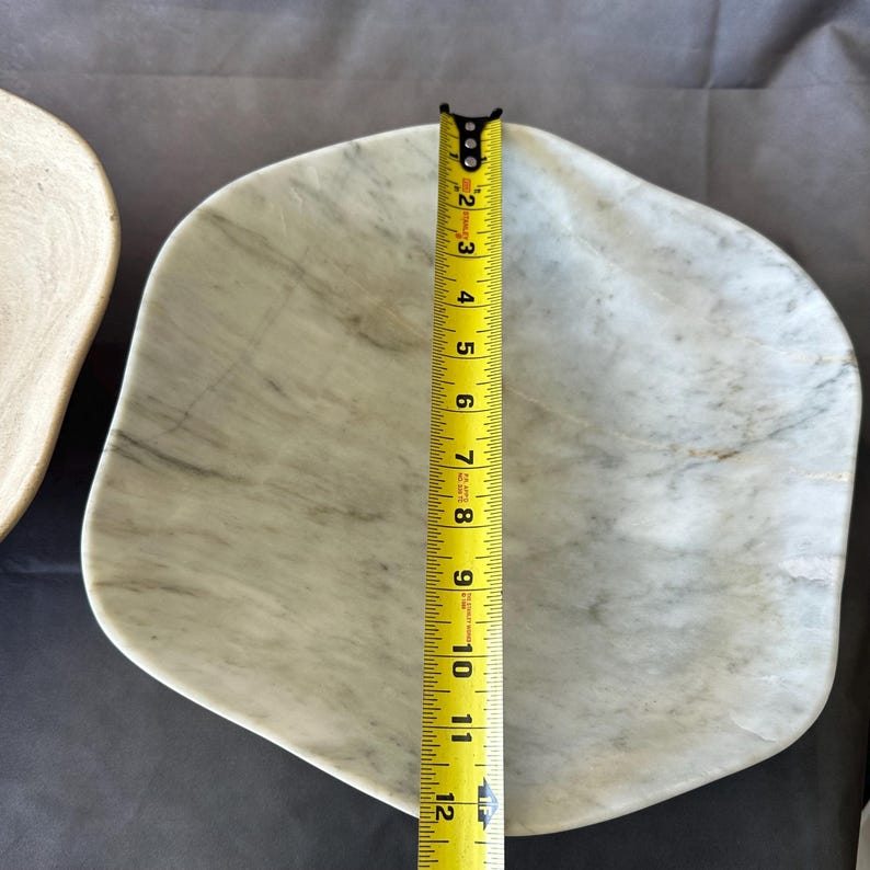 May include: A white marble serving dish with gray veining. The dish is irregularly shaped and measures approximately 11 inches in length. A yellow measuring tape is placed next to the dish to show its size.