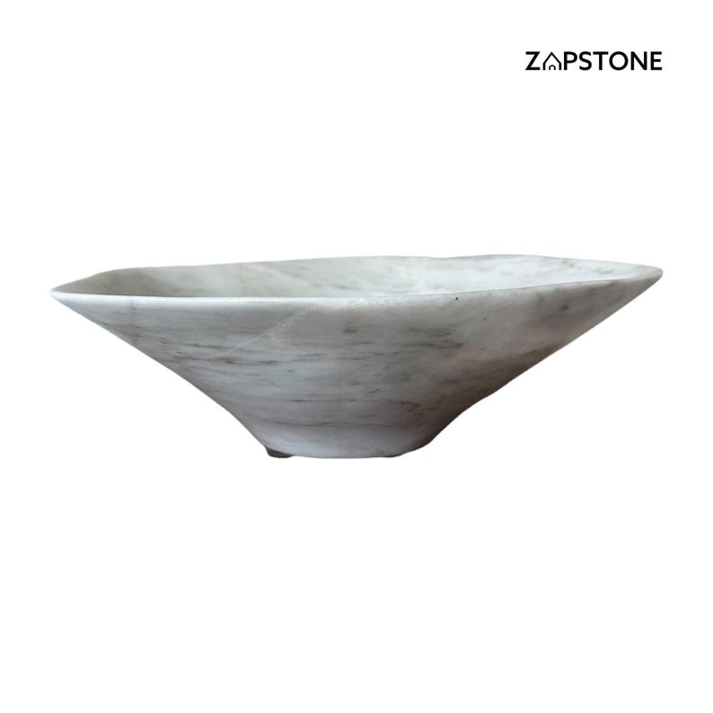 May include: A white marble bowl with a unique, asymmetrical shape. The bowl has a wide, open top and tapers down to a narrow base. The marble has subtle gray veining throughout. The brand name "ZAPSTONE" is printed in black on the upper right.