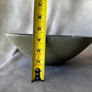 May include: A gray, stone-like bowl with a tape measure showing a height of approximately 3 inches. The bowl has a smooth surface and a slightly irregular shape. The tape measure is yellow with black markings.