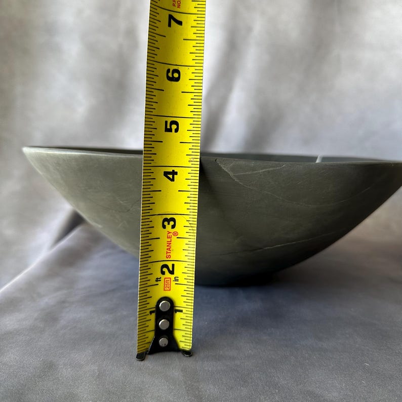 May include: A gray, stone-like bowl with a tape measure showing a height of approximately 3 inches. The bowl has a smooth surface and a slightly irregular shape. The tape measure is yellow with black markings.