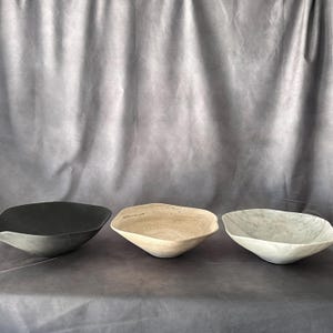 May include: Three irregularly shaped bowls in a row. The bowls are in shades of black, beige, and gray. The bowls have a textured surface and a shallow, open design. The background is a gray fabric.