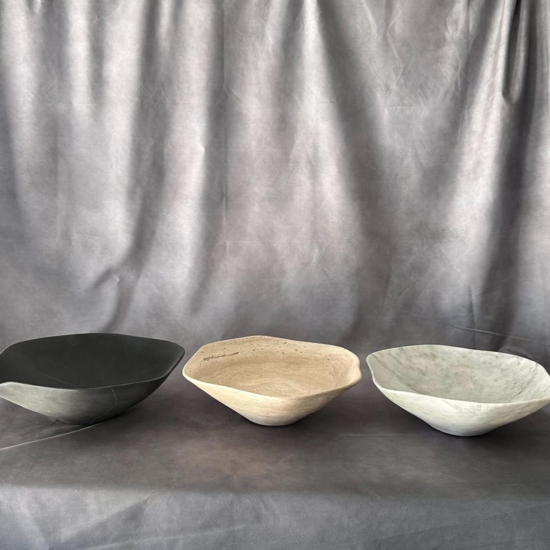 May include: Three irregularly shaped bowls in a row. The bowls are in shades of black, beige, and gray. The bowls have a textured surface and a shallow, open design. The background is a gray fabric.