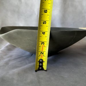 May include: A dark gray, square-shaped bowl with a wavy rim. The bowl is approximately 2 inches (5 cm) deep, as measured by a yellow measuring tape. The bowl's interior is not visible.