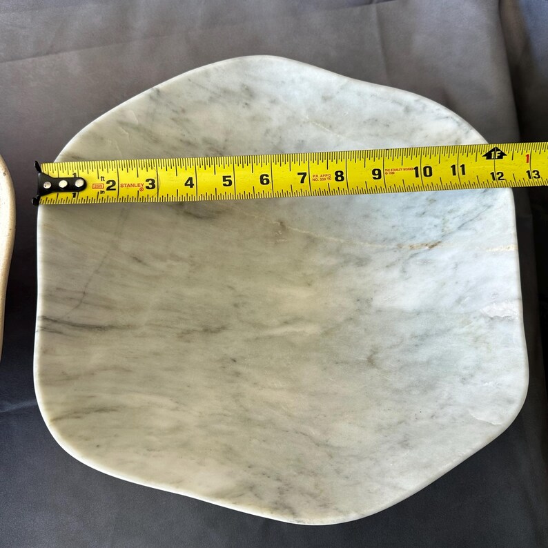 May include: A light gray marble serving platter with a unique, irregular shape. The platter is approximately 12 inches long. A yellow tape measure is placed across the platter to show its length.