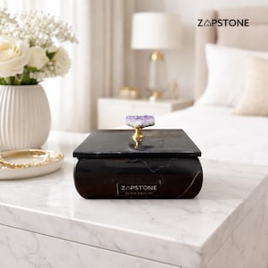 May include: A black marble box with a purple and gold gemstone handle. The box is rectangular with rounded edges and sits on a white marble surface. The text "ZAPSTONE HOME DECOR" is printed on the front of the box.