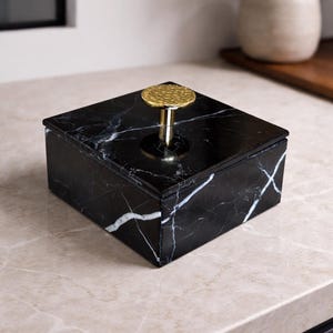 May include: A black marble box with white veining and a gold-colored, textured knob. The square box is on a light-colored surface. The lid has a shiny, reflective surface. A decorative storage box.