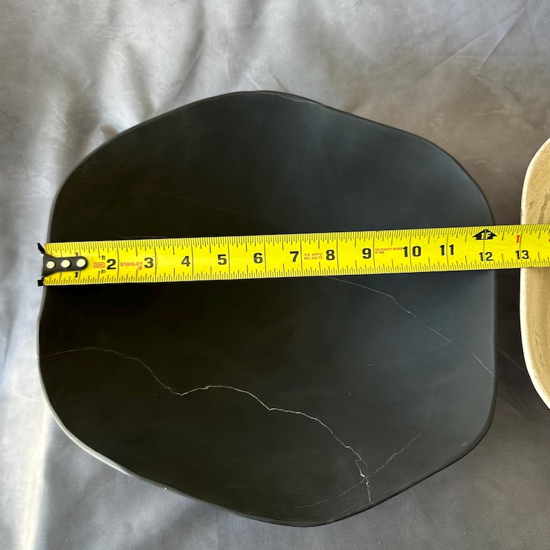 May include: A black, irregularly shaped plate with a white vein-like pattern. A yellow tape measure shows the plate is approximately 12 inches long. The plate is likely made of ceramic or stone.